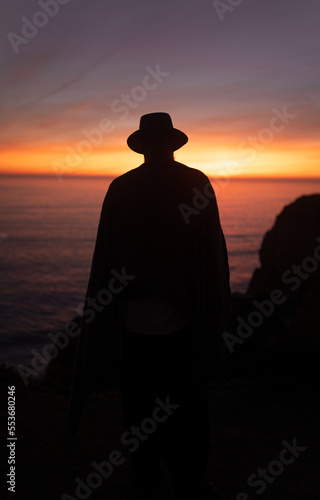 Silhouette of a man watching the ocean during the sunset wearing a poncho and a hat