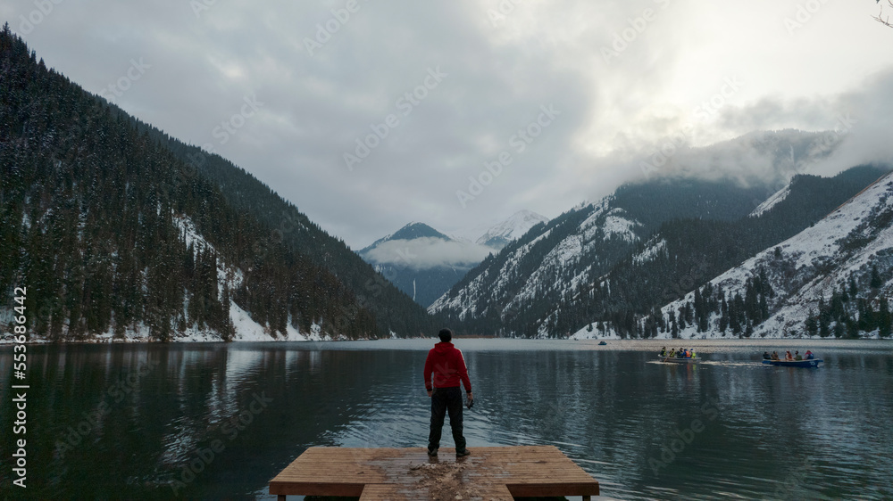 The guy standing on the pier admires the mountain lake. There is a view of the mirrored black color of the water, which reflects snowy mountains, green forest, clouds and a yellow sunset. Kolsai Lake