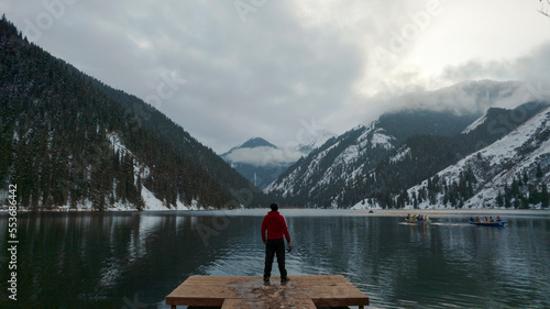 The guy standing on the pier admires the mountain lake. There is a view of the mirrored black color of the water, which reflects snowy mountains, green forest, clouds and a yellow sunset. Kolsai Lake