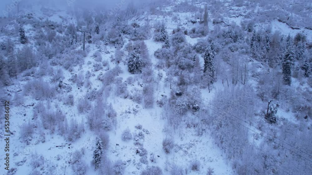 Cable car in the mountains in the winter forest. The cabins move along the gondola road through the winter snow forest and fog. White clouds covered the mountains. Top view from a drone. Medeo, Almaty Cable car in the mountains in the winter forest. The cabins move along the gondola road through the winter snow forest and fog. White clouds covered the mountains. Top view from a drone. Medeo, Almaty
