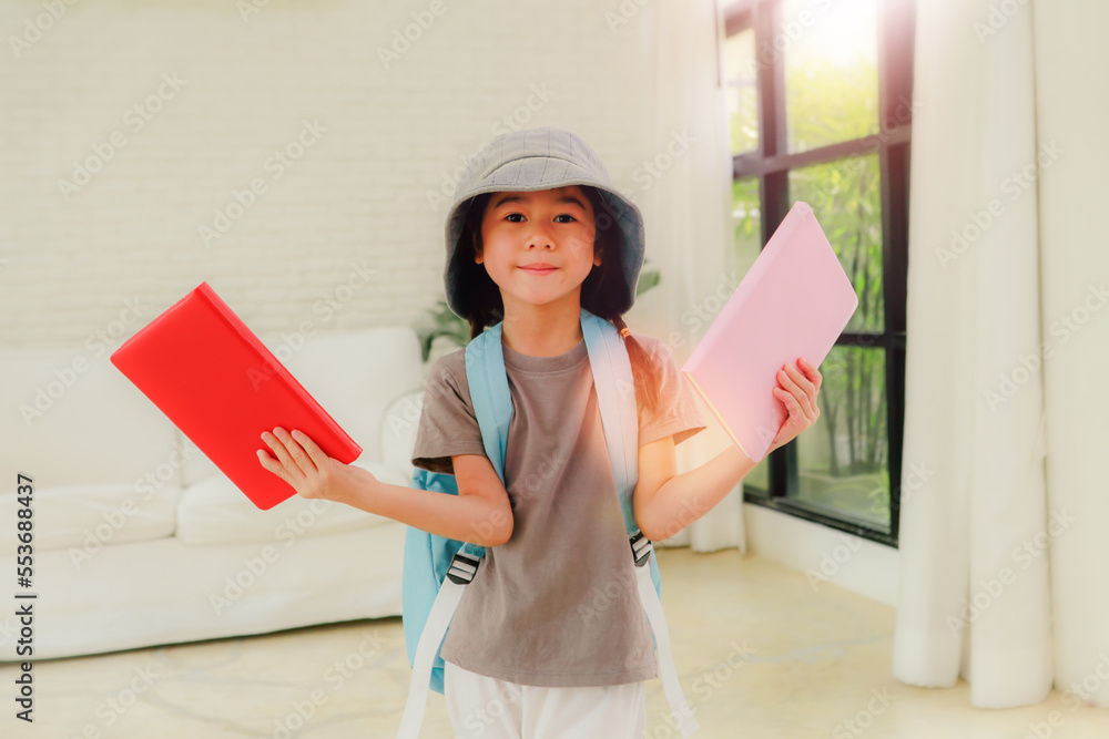 Portrait cute little Asian girl wearing a hat and a backpack holds two teaching books to do her homework after coming home from elementary school with a happy face. Portrait cute little Asian girl wearing a hat and a backpack holds two teaching books to do her homework after coming home from elementary school with a happy face.