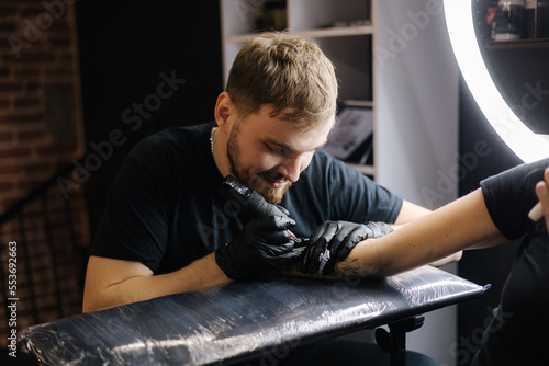 Handsome tattoo master making black tattoo with paint. Master works in black sterile gloves. Young tattoo artist in work