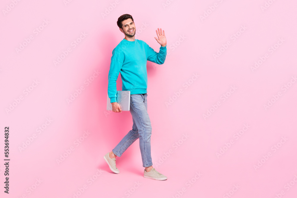 Full body photo of handsome young man hold netbook walking wave arm dressed stylish blue garment isolated on pink color background Full body photo of handsome young man hold netbook walking wave arm dressed stylish blue garment isolated on pink color background