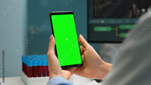 Close up of doctor's hand holding phone with green screen sitting at desk in biological laboratory while nurse bringing blood samples. Scientist using smartphone with mockup, chroma key display