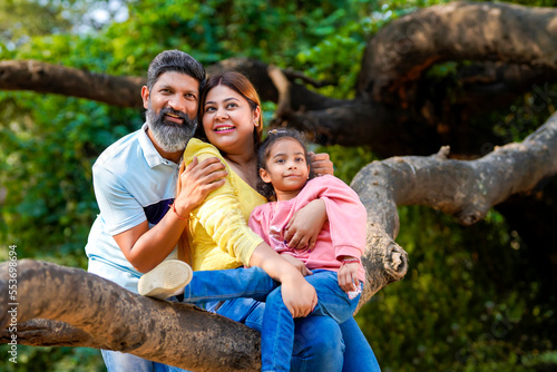 Happy indian couple sitting with his little girl on tree branch at garden.