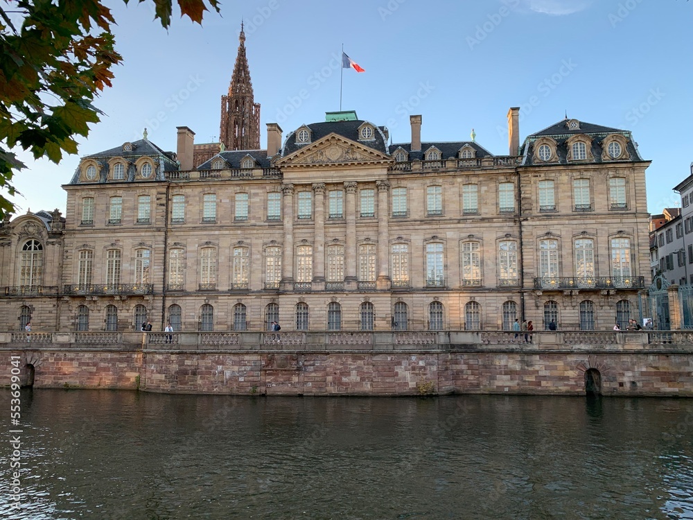 French flag is waving on top of baroque style Rohan palace (Palais Rohan). View from waterfront, river, canal side. Strasbourg cathedral tower in the background. Strasbourg, France