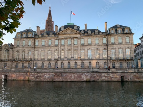 French flag is waving on top of baroque style Rohan palace (Palais Rohan). View from waterfront, river, canal side. Strasbourg cathedral tower in the background. Strasbourg, France