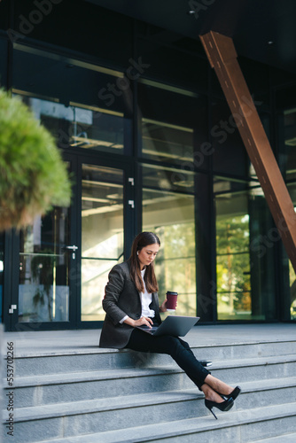 Beautiful young woman wearing jacket using laptop while sitting on stairs, drinking takeaway coffee cup. Modern communication.