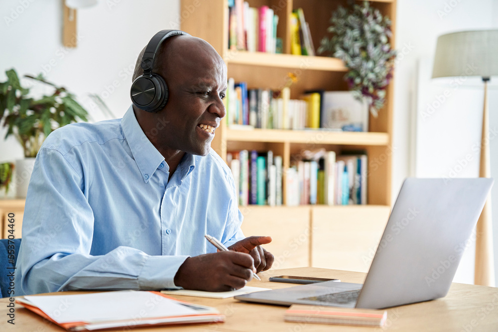 Obraz premium Middle-aged African-American man wearing headphones and taking notes while attending remote conference call while working at home with laptop computer