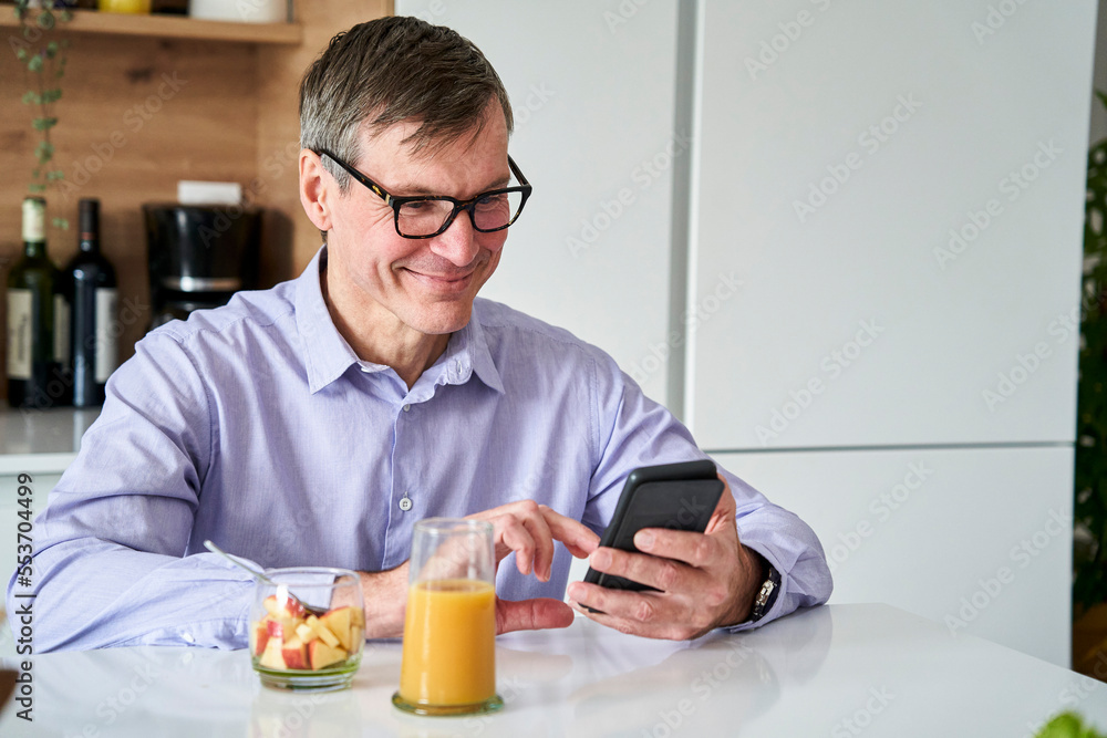Middle-aged professional man smiling and checking messages on phone while having breakfast on kitchen counter at home