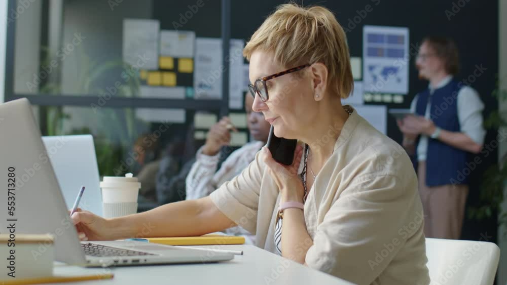 Businesswoman speaking on mobile phone, using laptop and taking notes during workday in office Businesswoman speaking on mobile phone, using laptop and taking notes during workday in office