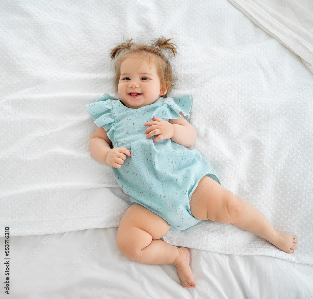 Happy baby girl smiling and looking at camera. Cute child with pigtails and blue suit pyjama in white bedroom lying on bed. Childhood, babyhood, people concept. Happy baby girl smiling and looking at camera. Cute child with pigtails and blue suit pyjama in white bedroom lying on bed. Childhood, babyhood, people concept.
