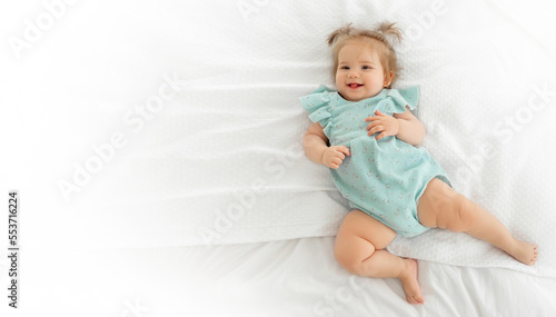 Happy baby girl smiling and looking at camera. Cute child with pigtails and blue suit pyjama in white bedroom lying on bed. Childhood, babyhood, people concept. Copy space