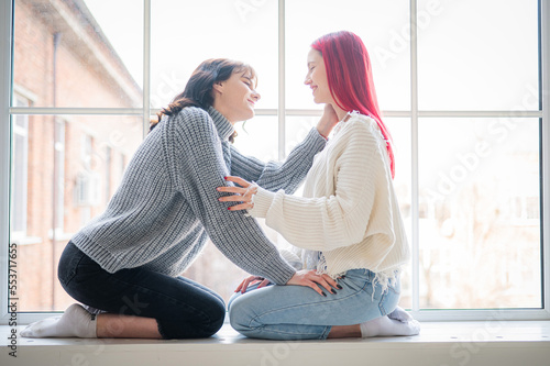 Two women dressed in sweaters sit by the window and gently hug. Lesbian intimacy.