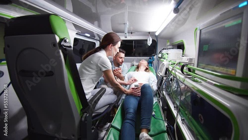 Professional female paramedic listening to heartbeat of baby with stethoscope providing help for young pregnant woman suffering from labor contractions in ambulance. Husband supporting wife. photo