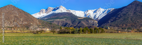 Mountain panorama with snow-capped peaks, in a valley with green meadows, and a village with a small castle. We see a small chapel and old medieval towers, built high on the mountain. Alps, France.