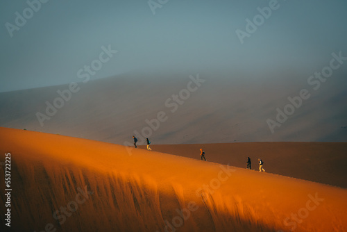 Touristen wandern über einen von der Morgensonne erleuchteten Grat in Richtung des nebelverhangenen Gipfels der Düne Big Daddy im Sossusvlei (Namibia) photo