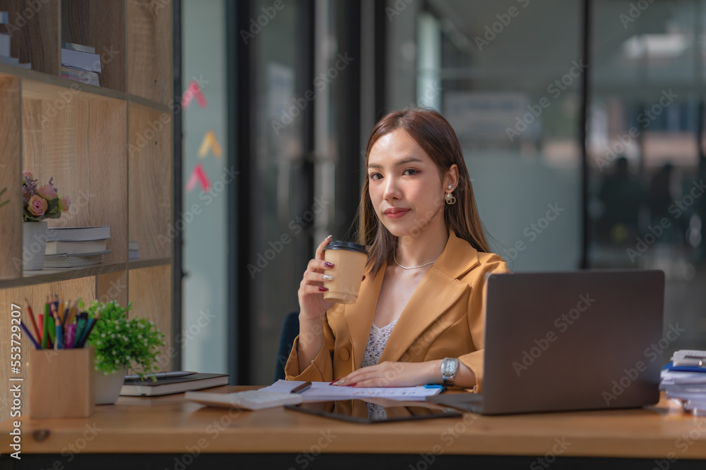Beautiful woman holding a coffee cup and using a laptop while sitting at her working place in an office. Beautiful woman holding a coffee cup and using a laptop while sitting at her working place in an office.