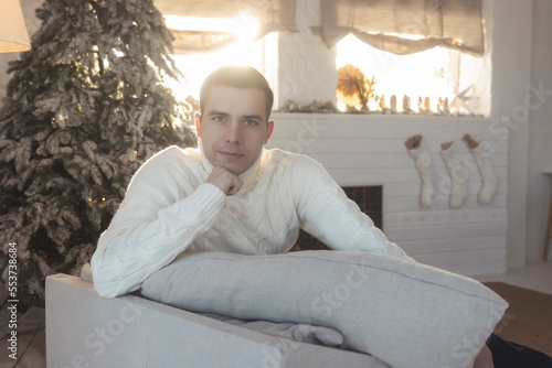 a man in a white sweater against the background of New Year's decorations sits in an armchair © drakuliren