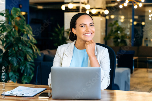 Beautiful positive clever elegant brazilian or hispanic young woman, secretary, manager, financial director, sitting at a desk with laptop in a modern office, looking at the camera, smiling friendly