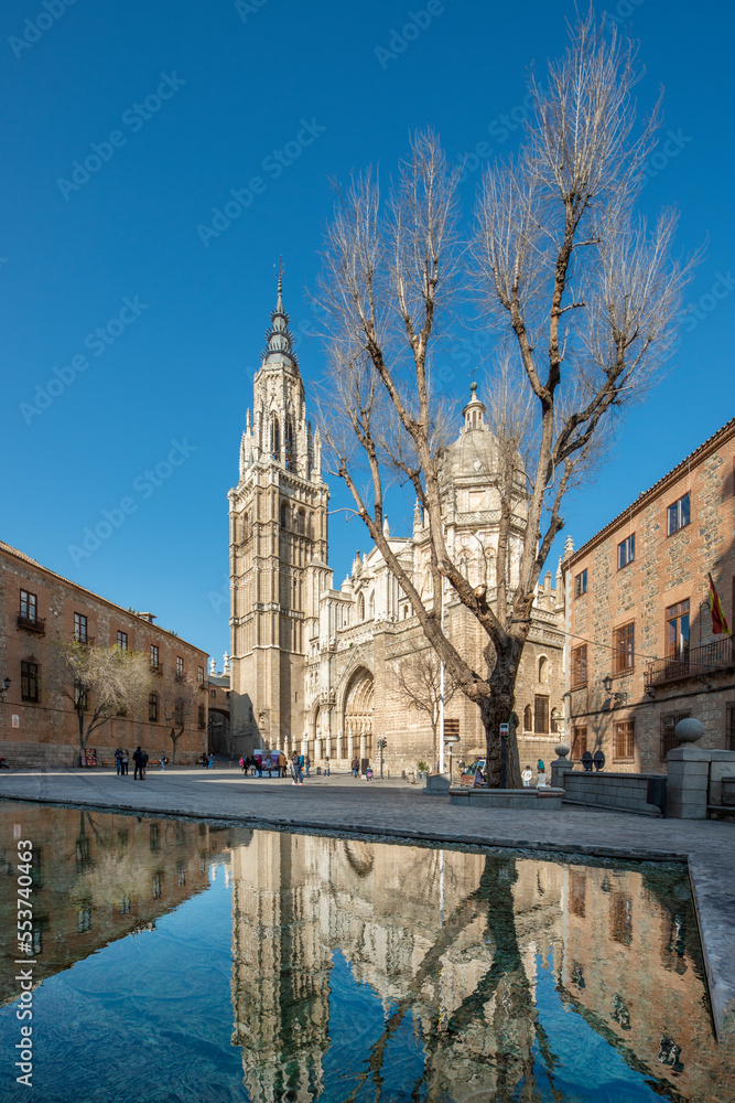 Facade of the cathedral of Toledo, Spain and a large tree with reflection in the water of a decorative fountain on a winter day with clear skies