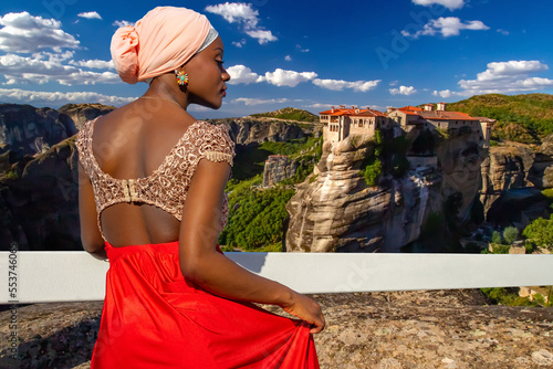 African American woman admires nature. Girl looks at rock building. Woman travels in nature. Woman leans on white fence. Girl tourist with her back to camera. Meteora in Greece. Varlaam monastery photo