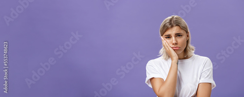 Waist-up shot of unhappy miserable and sad cute blonde female in white casual t-shirt pursing lips leaning face on palm and frowning from disappointement and regret over purple background photo