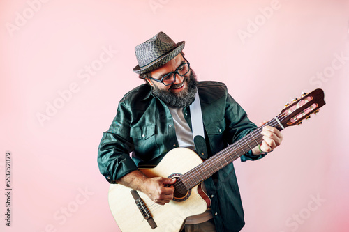 Happy bearded man playing acoustic guitar on pink background.