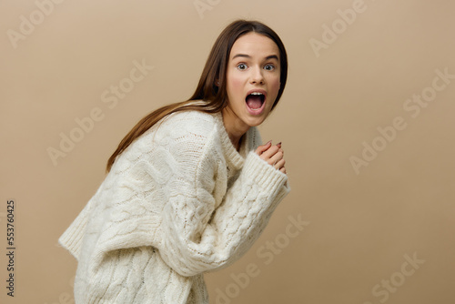 the surprised woman is standing half sideways and looking straight at a light brown background in a white sweater leaned forward pressing her hand to herself © Tatiana