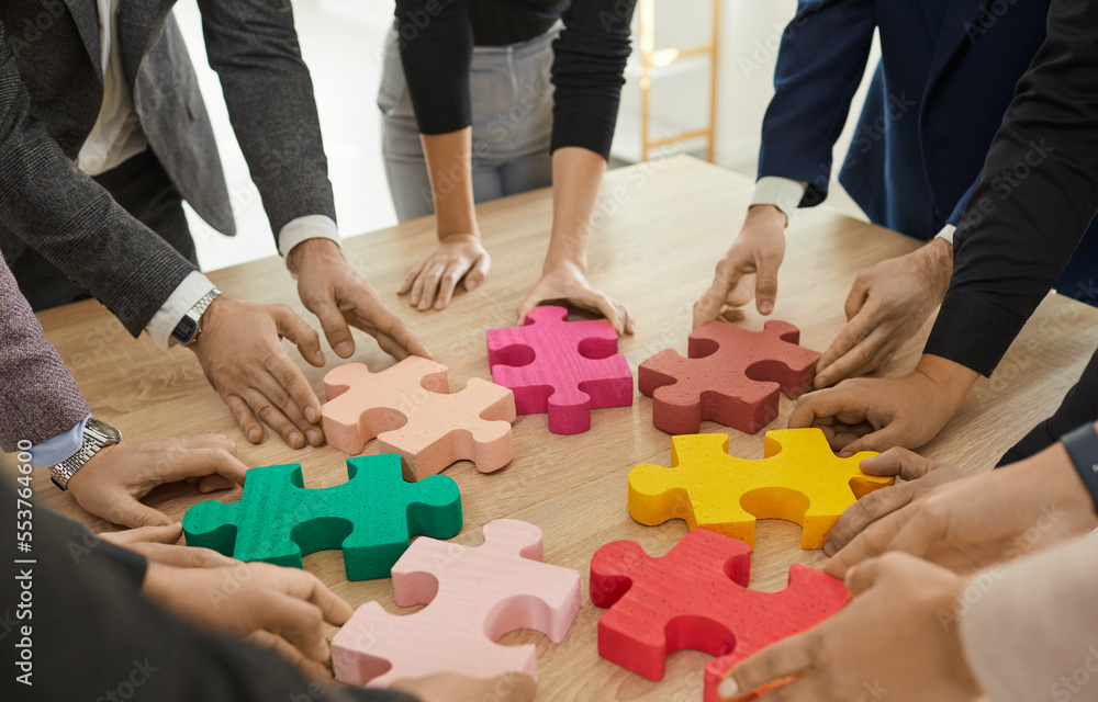 Business team trying to match pieces of jigsaw puzzle. Group of young people standing around wooden table and connecting colorful red, yellow, green, pink jigsaw parts. Cropped shot. Teamwork concept Business team trying to match pieces of jigsaw puzzle. Group of young people standing around wooden table and connecting colorful red, yellow, green, pink jigsaw parts. Cropped shot. Teamwork concept
