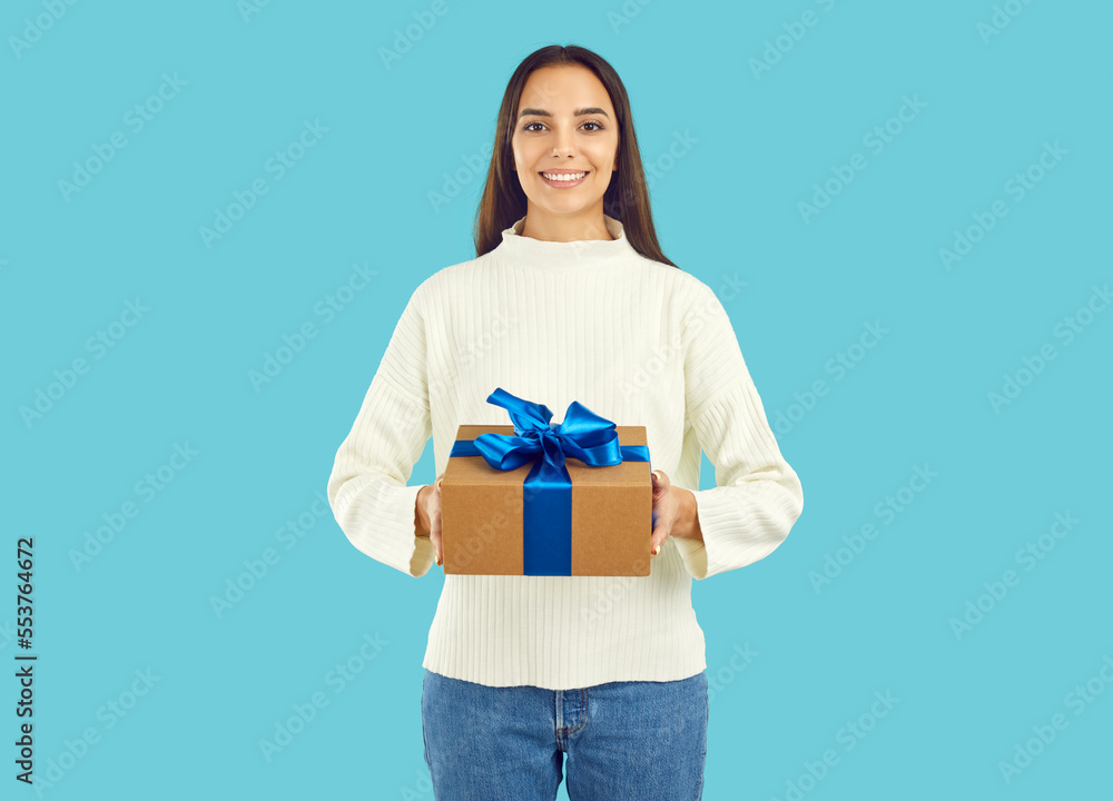 Studio portrait of happy attractive young brunette woman in warm white winter sweater standing on blue color background holding gift box with Christmas present tied with beautiful blue bow and smiling Studio portrait of happy attractive young brunette woman in warm white winter sweater standing on blue color background holding gift box with Christmas present tied with beautiful blue bow and smiling
