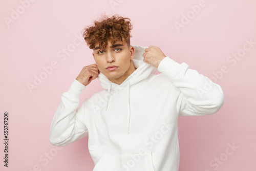 a handsome, attractive man stands on a pink background in a stylish white hoodie with a hood on his head and holds it with his hands, correcting it, and looks pleasantly into the camera