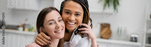 happy african american lesbian woman hugging joyful girlfriend in living room, banner.