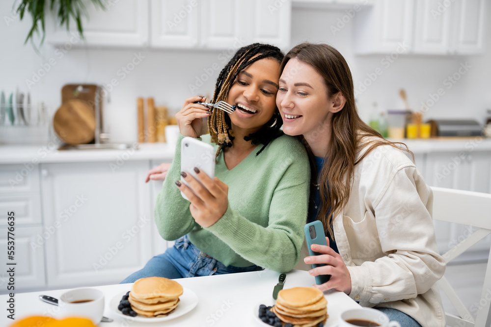 happy african american lesbian woman showing smartphone to girlfriend during breakfast.