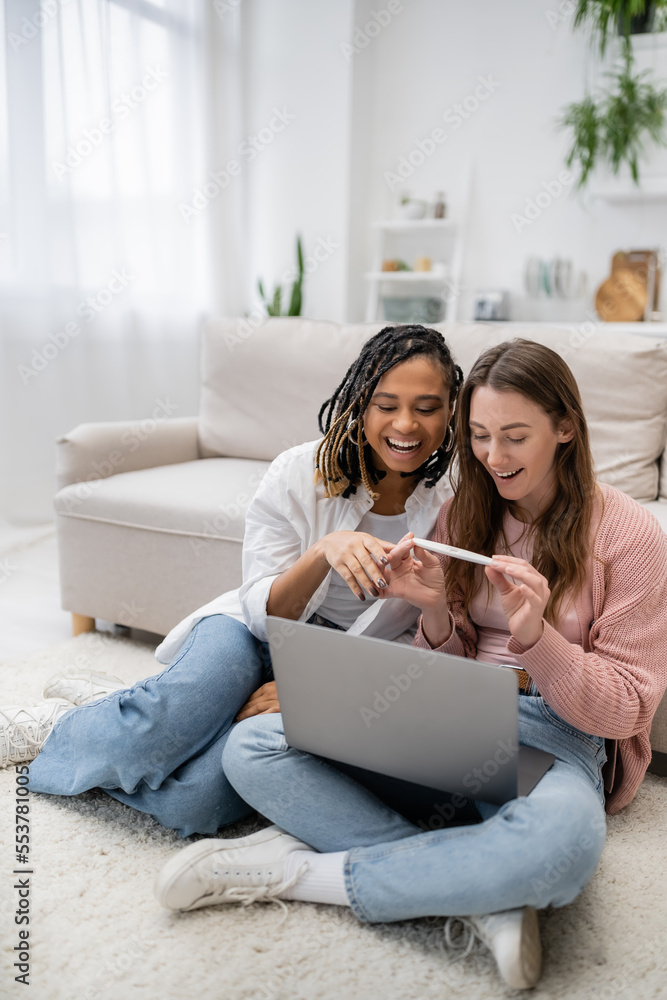 happy and lesbian woman showing pregnancy test near african american girlfriend during video call on laptop.