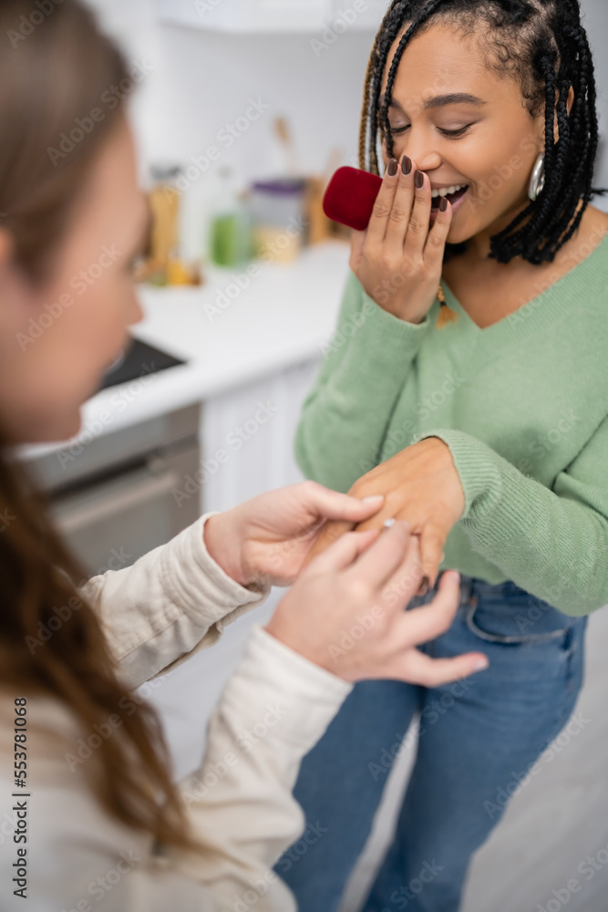 lesbian woman making proposal and wearing engagement ring on finger of amazed african american girlfriend. lesbian woman making proposal and wearing engagement ring on finger of amazed african american girlfriend.