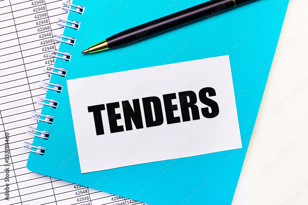 On the office desk there are reports, a blue notepad, a black pen, and a white card with the text TENDERS. Stylish workplace. Business concept On the office desk there are reports, a blue notepad, a black pen, and a white card with the text TENDERS. Stylish workplace. Business concept