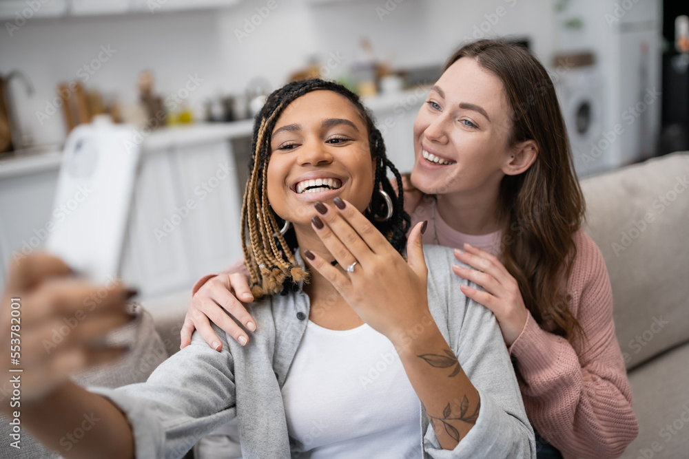 happy african american and lesbian woman showing engagement ring while taking selfie with smiling girlfriend.