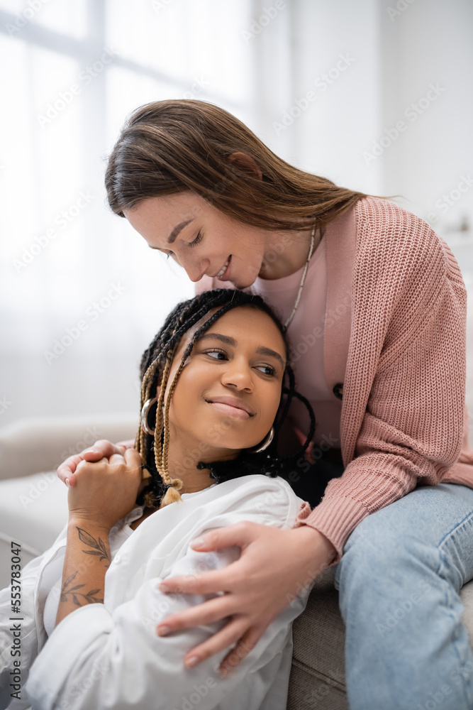 smiling lesbian woman sitting on couch and hugging tattooed african american girlfriend. smiling lesbian woman sitting on couch and hugging tattooed african american girlfriend.