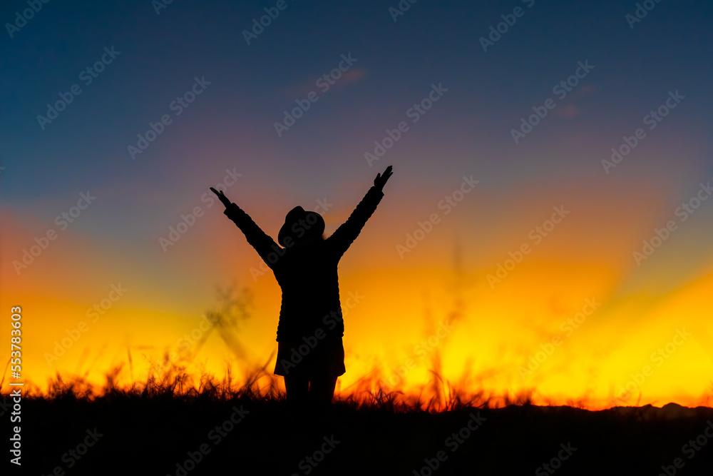 Silhouette of Christian woman raised hands and praying at meadow on sunset background. Hope, surrender and praising concept.