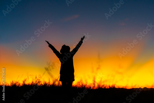 Silhouette of Christian woman raised hands and praying at meadow on sunset background. Hope  surrender and praising concept.