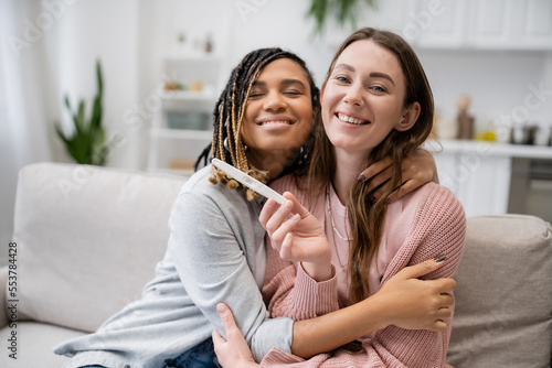 pleased african american and lesbian woman hugging happy girlfriend with pregnancy test.