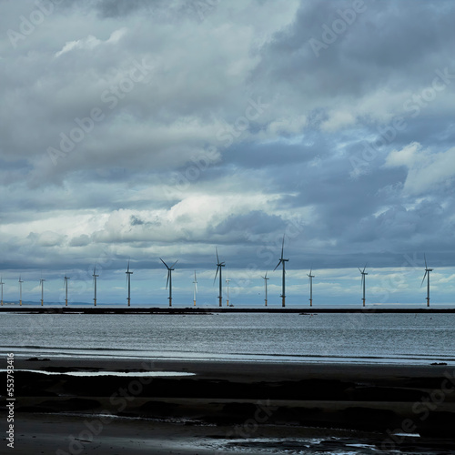 Cloud shadows play across a near-shore wind farm in North-East England, huge cumulus formations driven by the strong wind turning the massive turbines.