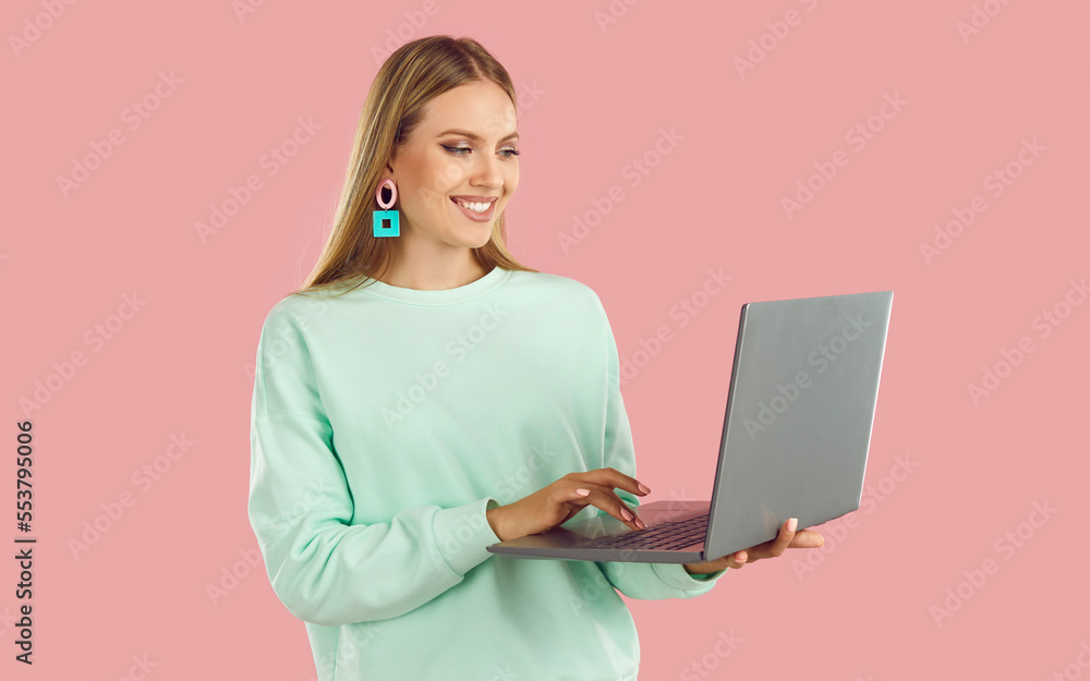 Happy blondy woman in mint sweatshirt doing remote job working on laptop on pink background and looking at screen. Distance work, online shopping, browsing social media, internet surfing concept. Happy blondy woman in mint sweatshirt doing remote job working on laptop on pink background and looking at screen. Distance work, online shopping, browsing social media, internet surfing concept.