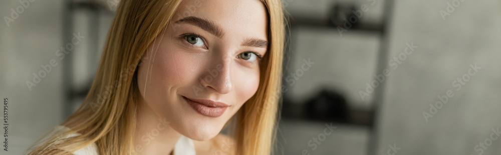 Young blonde woman smiling at camera in blurred living room, banner. Young blonde woman smiling at camera in blurred living room, banner.