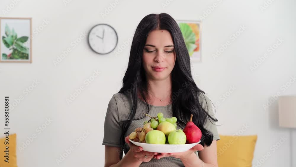nutrition during pregnancy, a young woman with plate of healthy tasty fruits in hands stand in room at home, smiling and looking at camera