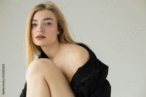 sensual blonde woman posing in black blazer and looking at camera isolated on grey.