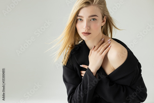Bild auf Leinwand blonde and seductive woman in black blazer touching bare shoulder while looking at camera isolated on grey