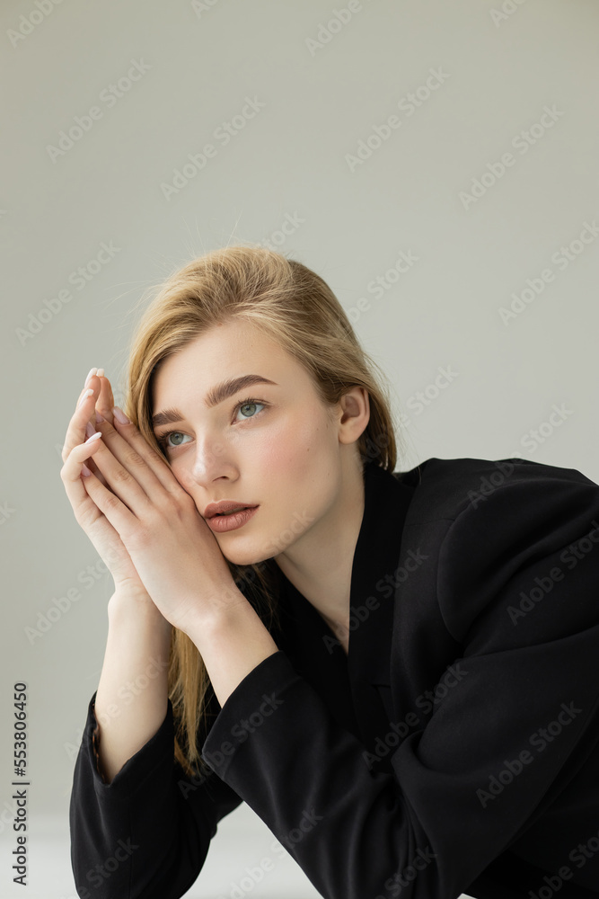 dreamy woman in black jacket holding hands near face and looking away isolated on grey. dreamy woman in black jacket holding hands near face and looking away isolated on grey.