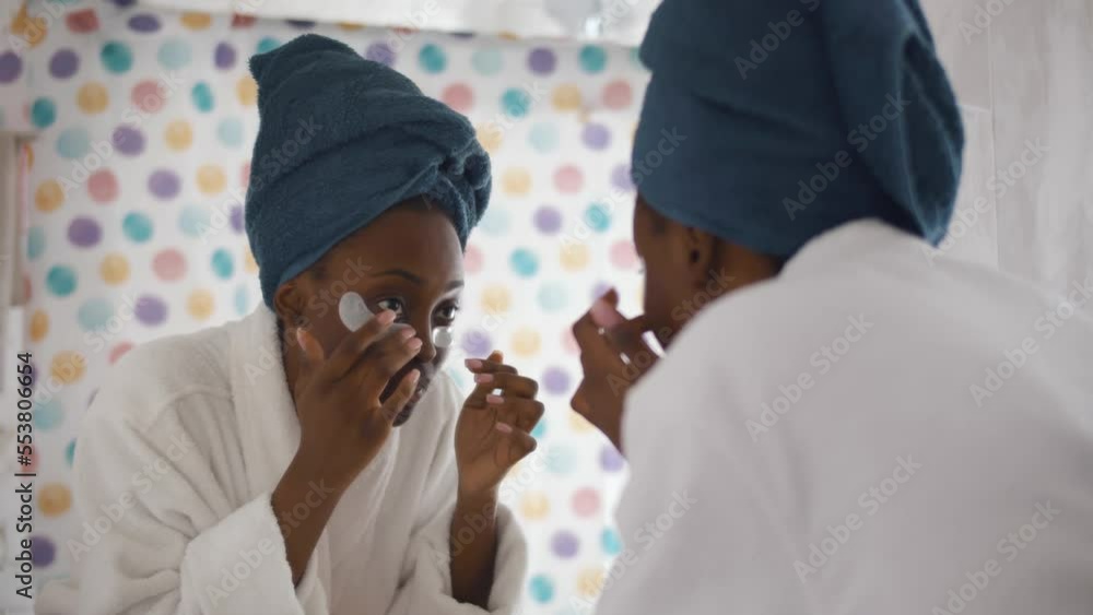 Happy African-American woman apply eye care patches looking in bathroom mirror. Realtime